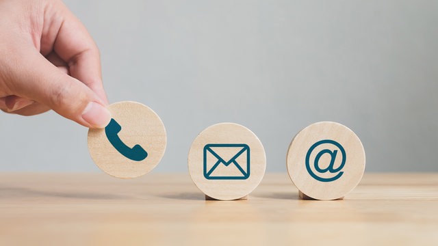 Eine Hand stellt mit runde Holzplättchen mit den Symbolen Telefon, E-Mail, Adresse auf (Symbolbild)